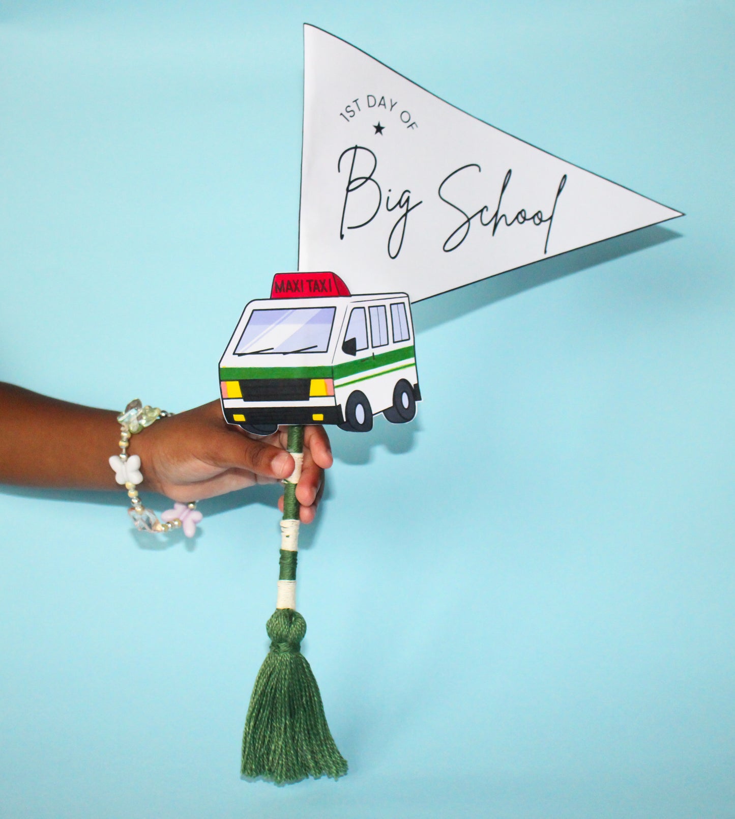 Hand holding a paper flag with a maxi taxi van illustration and '1st Day of Big School' text on a blue background