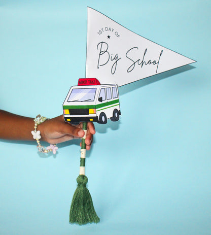 Hand holding a paper flag with a maxi taxi van illustration and '1st Day of Big School' text on a blue background