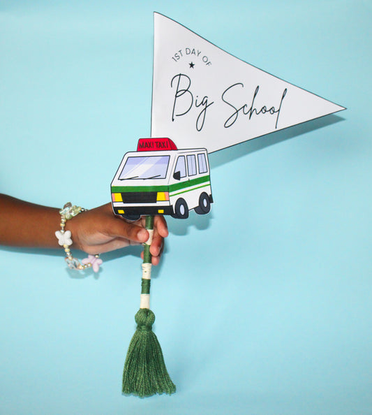 Hand holding a paper flag with a maxi taxi van illustration and '1st Day of Big School' text on a blue background