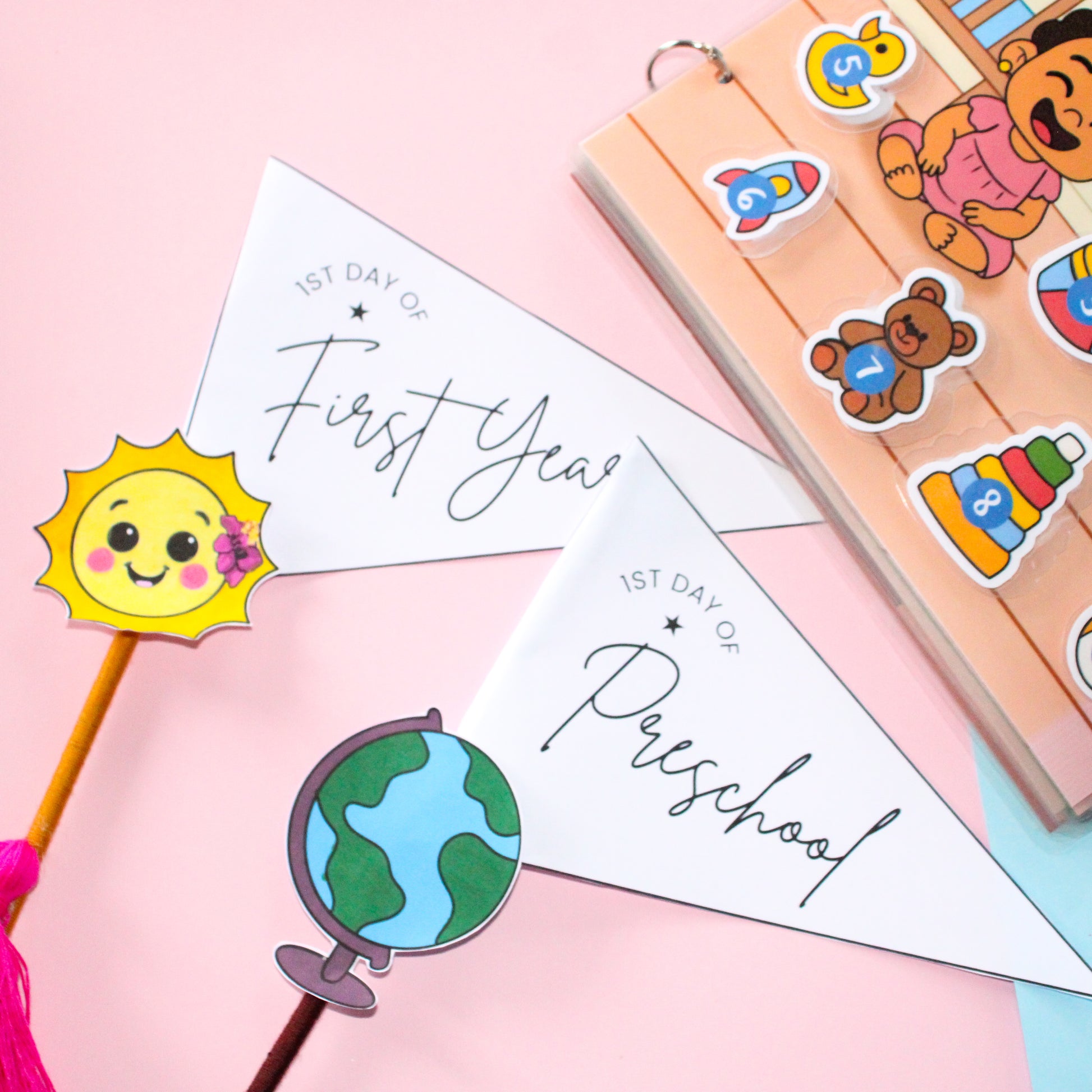 Children's school-themed items including a sun-shaped pennant flag, a globe-shaped pennant flag, and a busy book with removable velcro pieces on a pink background.