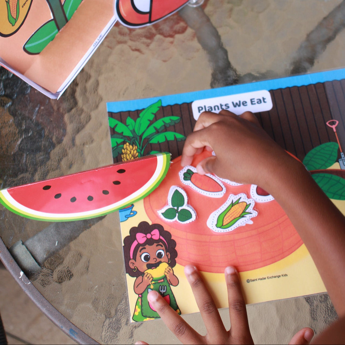 A child's hands are shown interacting with a colorful educational children's book or activity sheet about plants and foods, featuring stickers and cutout pieces on a glass table.